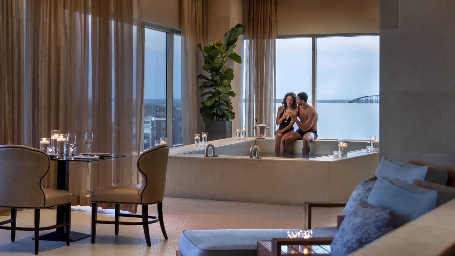 A couple enjoying a candlelit hot tub in a luxury hotel spa, with elegant seating, soft lighting and large windows offering a view of a bridge in the distance