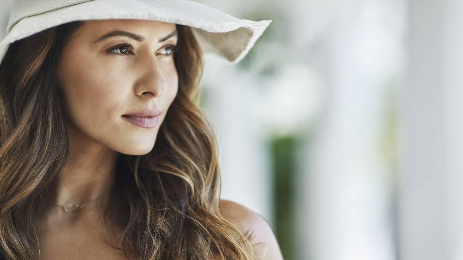 A woman with wavy brown hair and a white floppy hat looks off into the distance