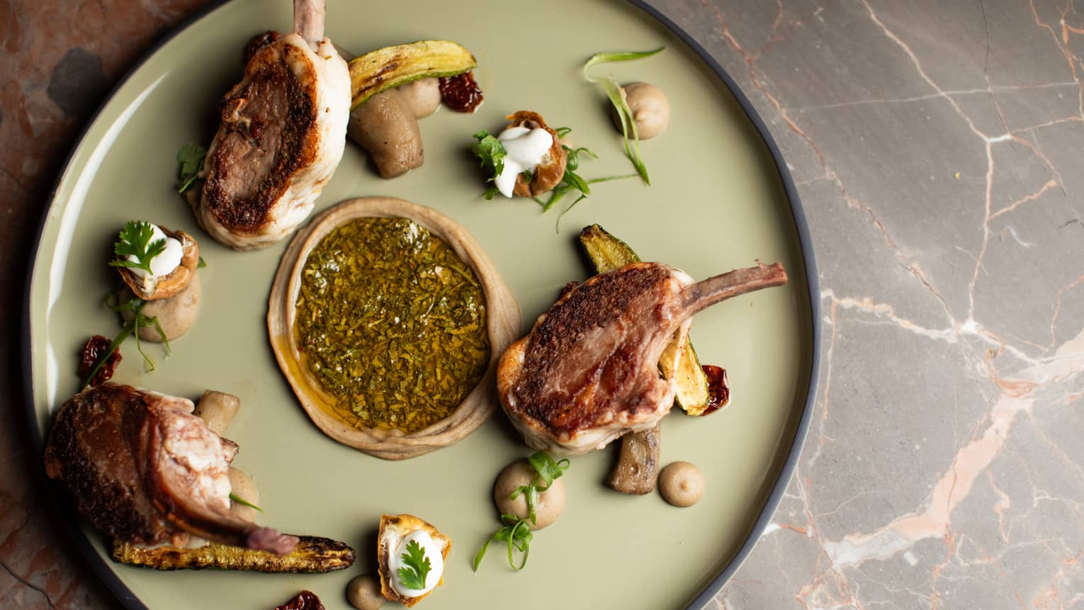 Three grilled lamb chops plated with eggplant and an assorment of garnishes on a large, round, light-green plate