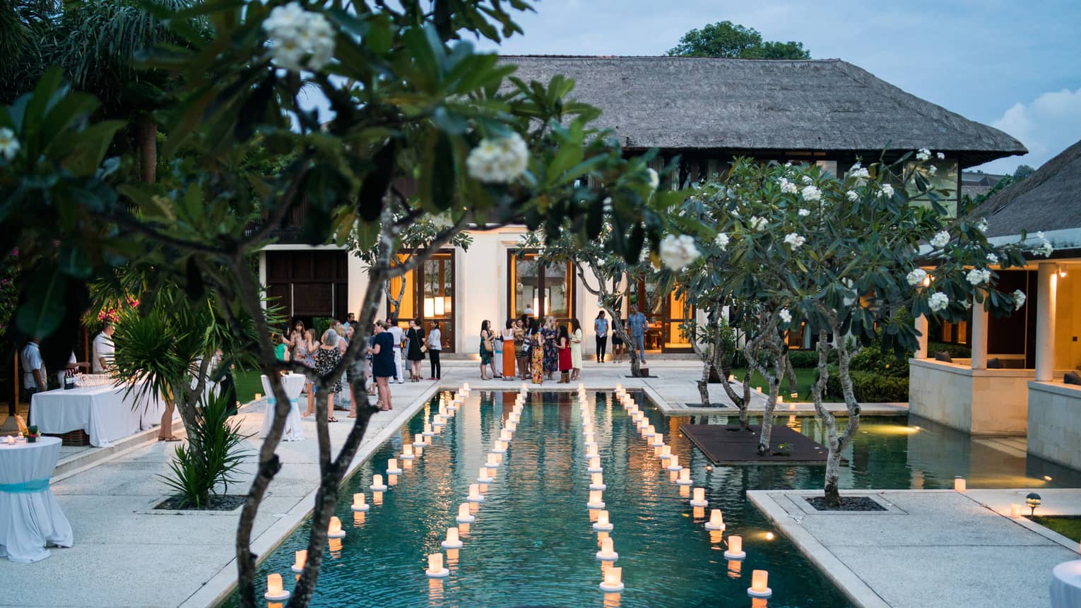 View past tropical tree to guests at patio reception around pool with floating candles