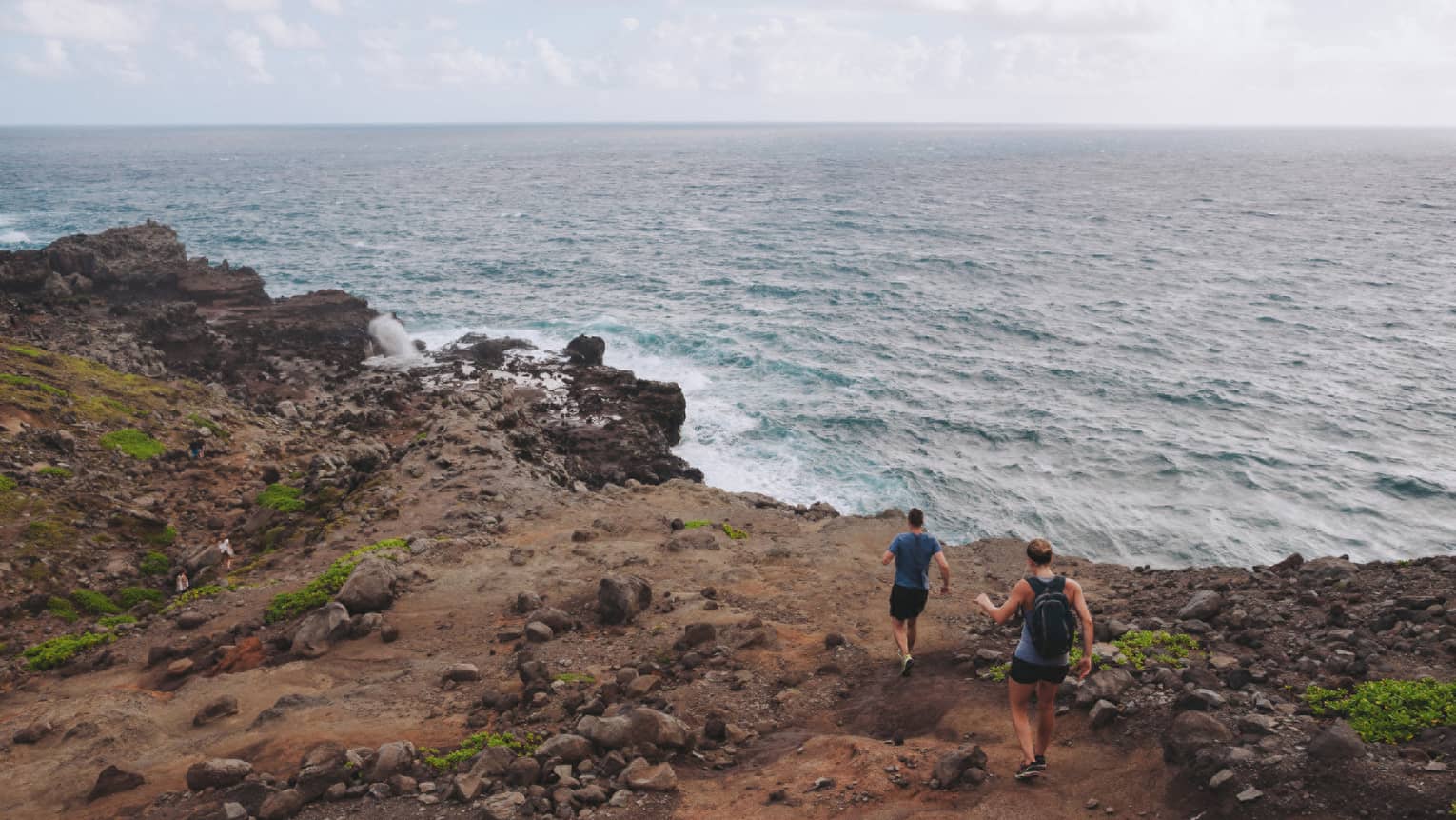 Two people hike down dirt-covered path, rocks towards ocean