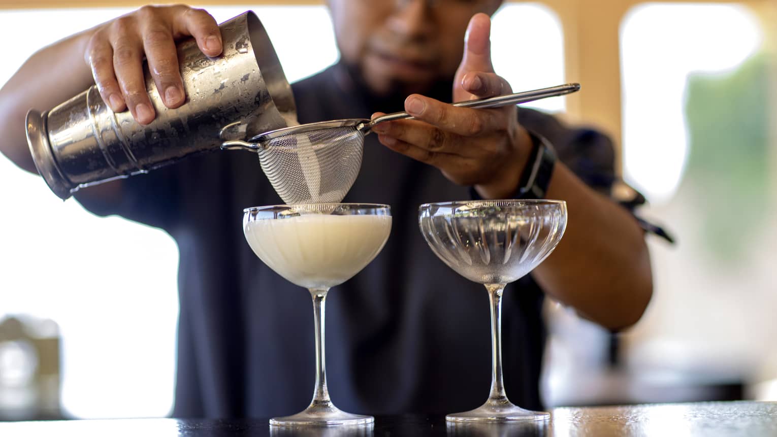 From a metal cocktail shaker, a bartender filters a milky cocktail through a sieve into an etched groove coupe glass. 