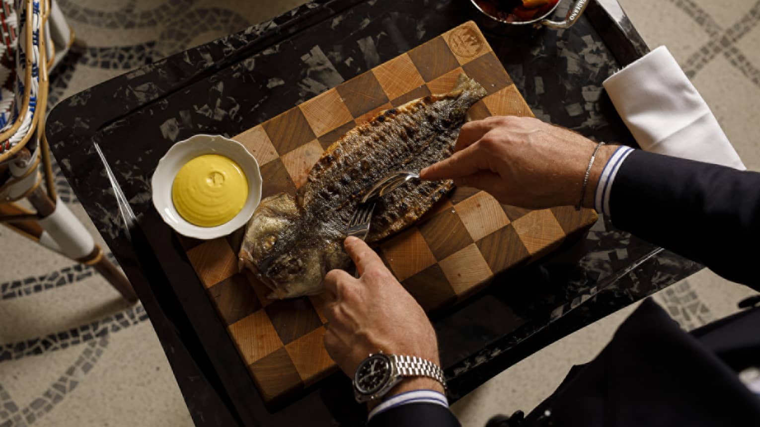 A person cuts into a plated cooked fish dish on a tray. Small bowls of condiments and sides are next to the serving plate on the tray.