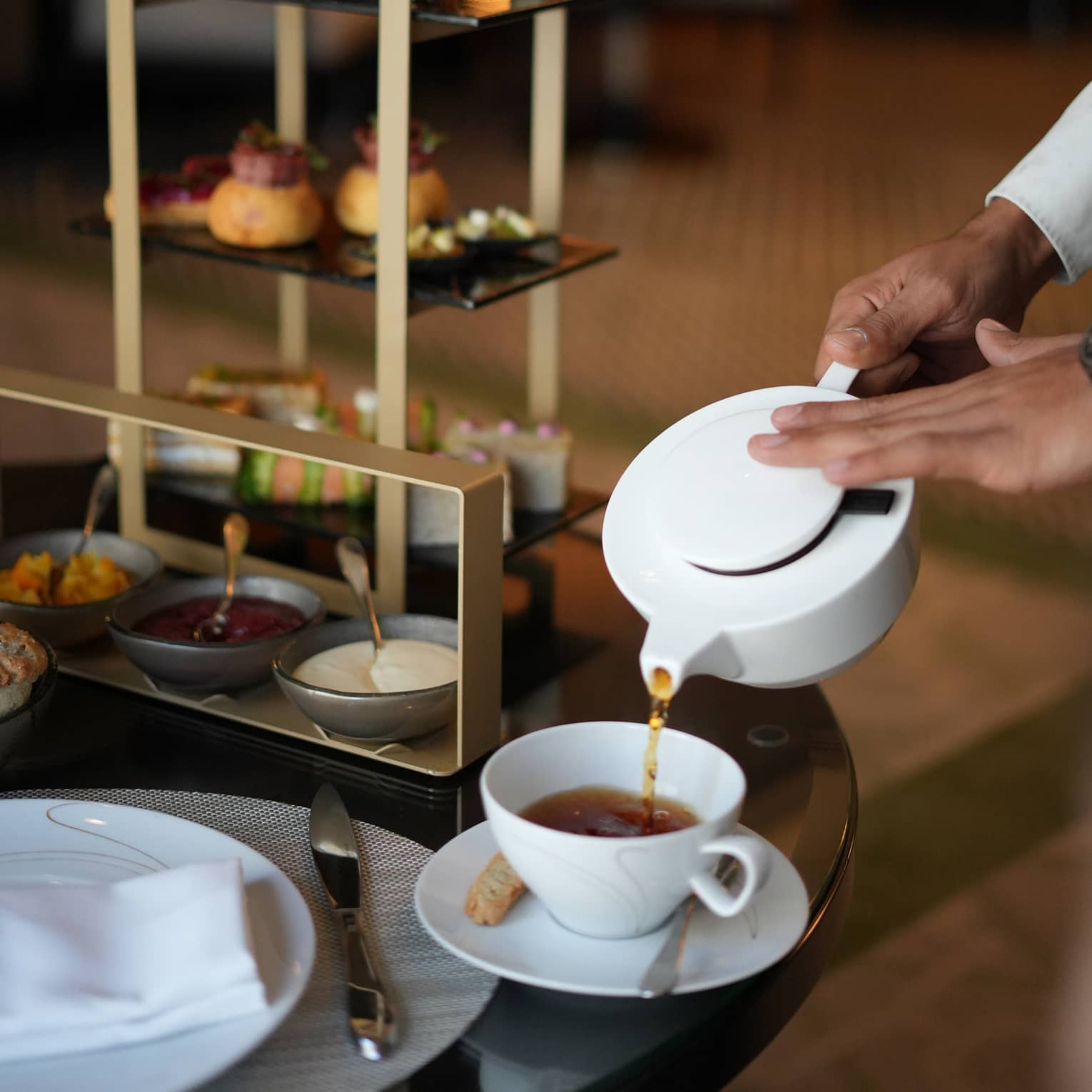 Hands pour tea from a white tea pot into a white mug set on a table next to a gold three-tiered tray filled with pastries for afternoon tea