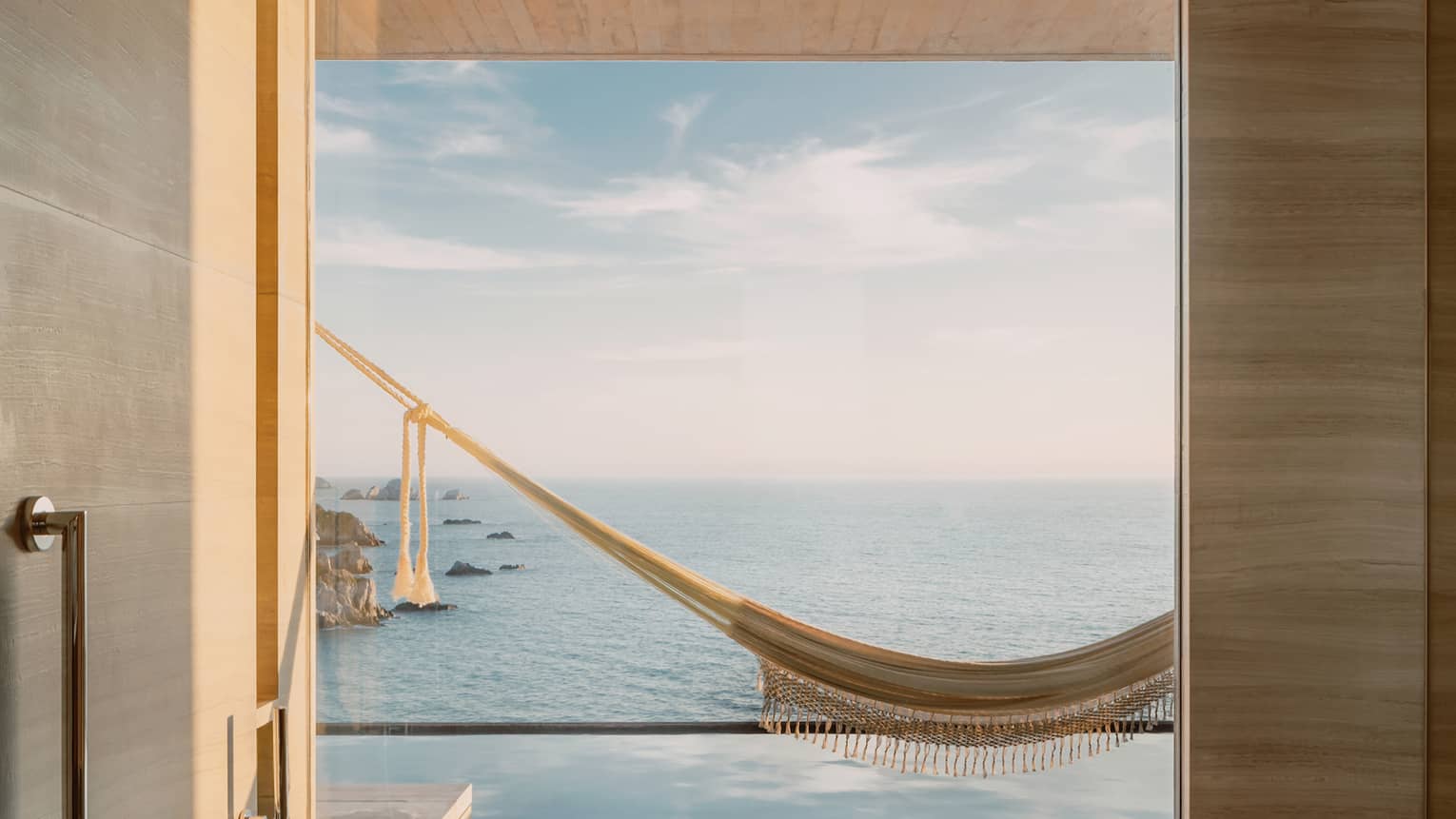 Minimalist bathroom with a deep tub and a large window offering a breathtaking view of an infinity pool, a hammock and the ocean stretching towards the horizon