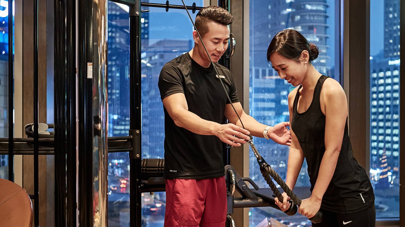 With skyscrapers out the window beyond, a trainer guides a guest as she performs a triceps pushdown exercise on a gym machine.
