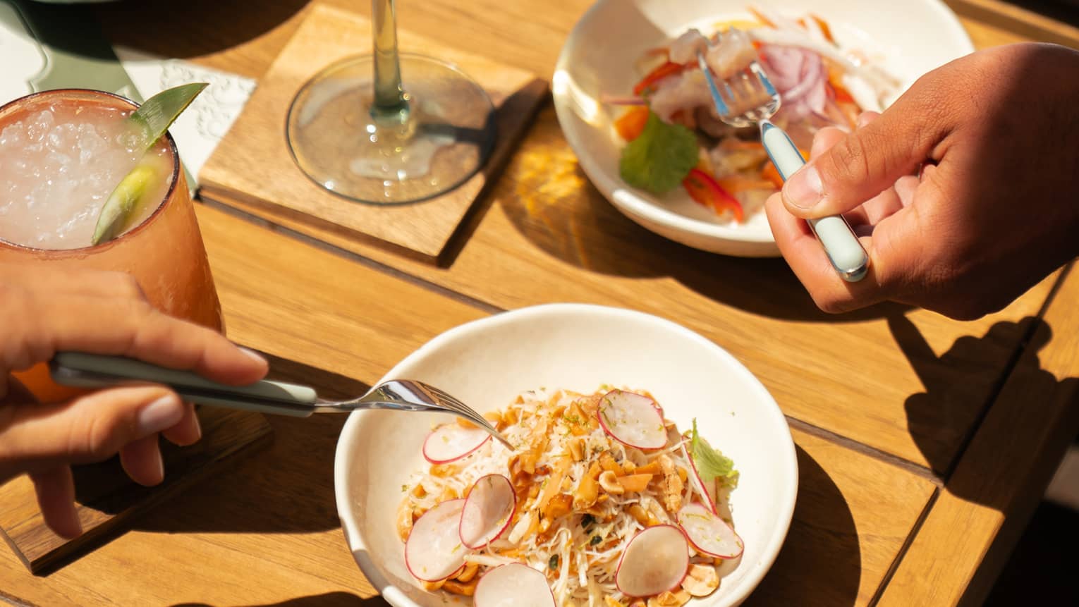 A wood table with small dishes of food items and cocktails in stemware and a tumbler. Two people reach for bites of food with forks.