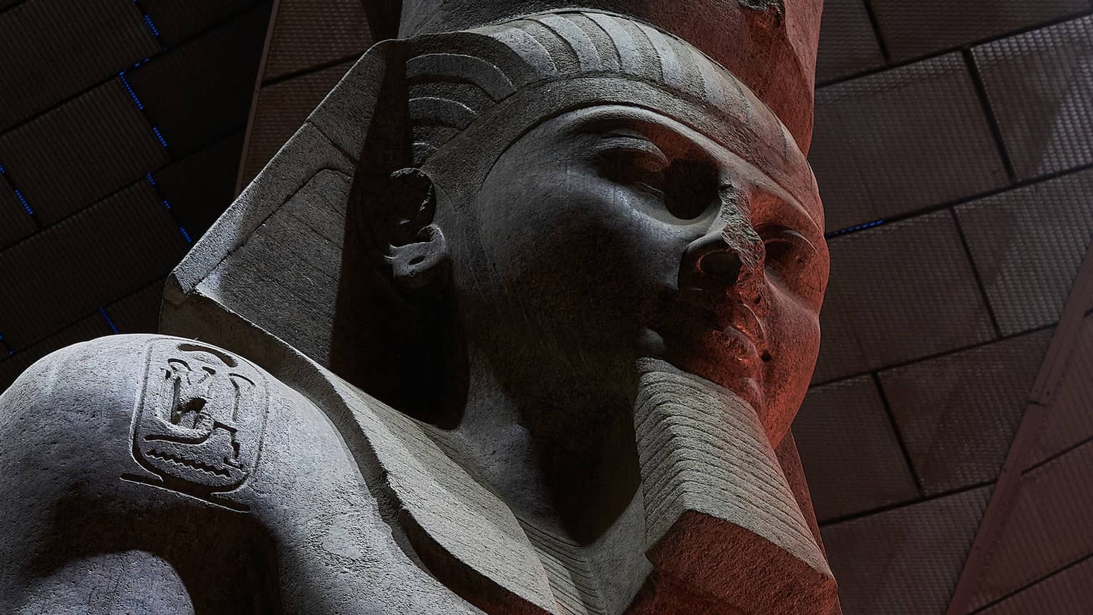 Close-up on a dark stone statue of a bearded pharoah with three heiroglyphic carvings on his chest and shoulders.