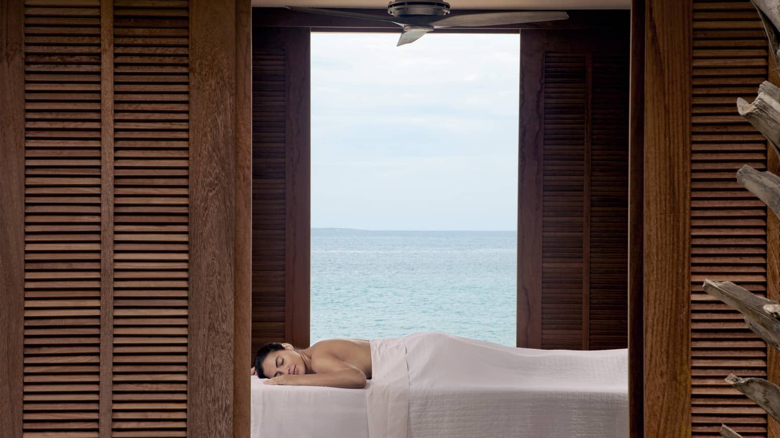 Guest enjoying a relaxing outdoor massage with a serene ocean view, framed by wooden shutters at a luxury beachfront resort