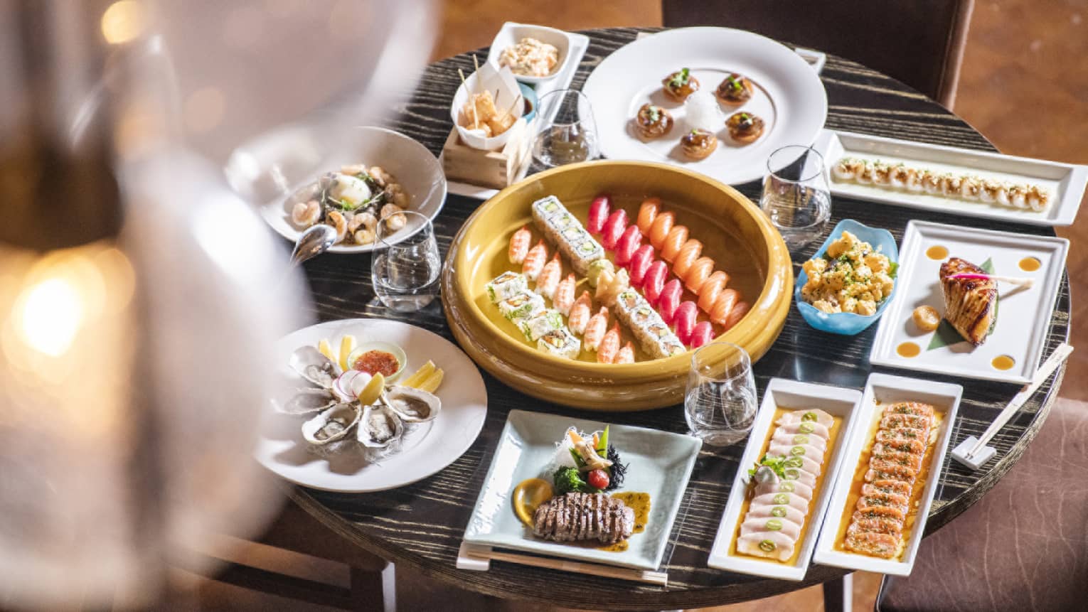 Small round table filled to the brim with a variety of square and round dishes filled with food, all surrounding a round gold platter with five rows of sushi in the center