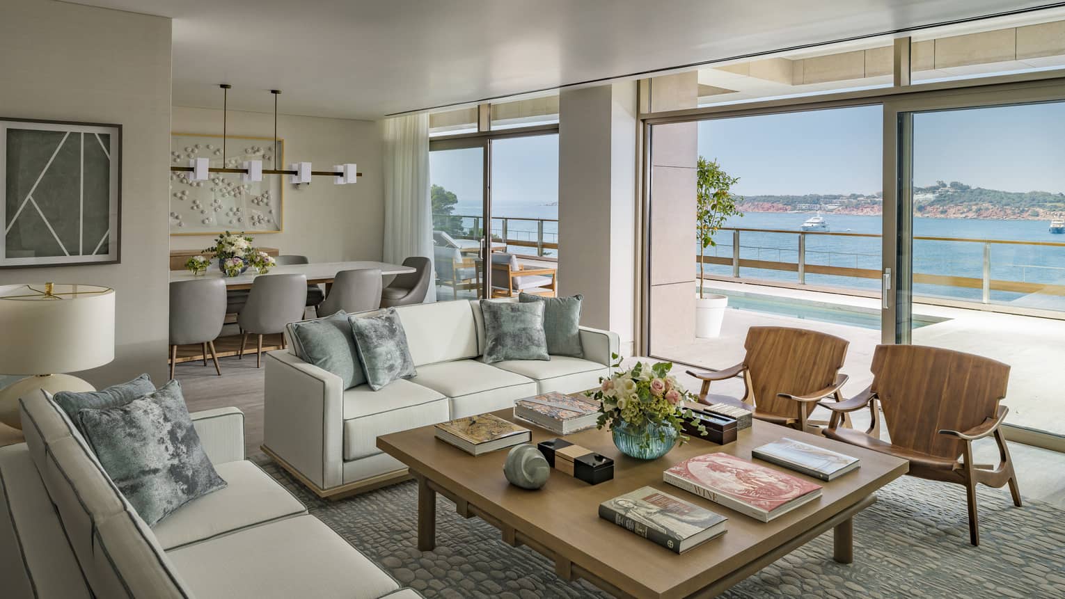 Arion Royal Suite living area with white sofas, square coffee table, arm chairs, dining table in background, floor-to-ceiling window with view of plunge pool and Mediterranean Sea