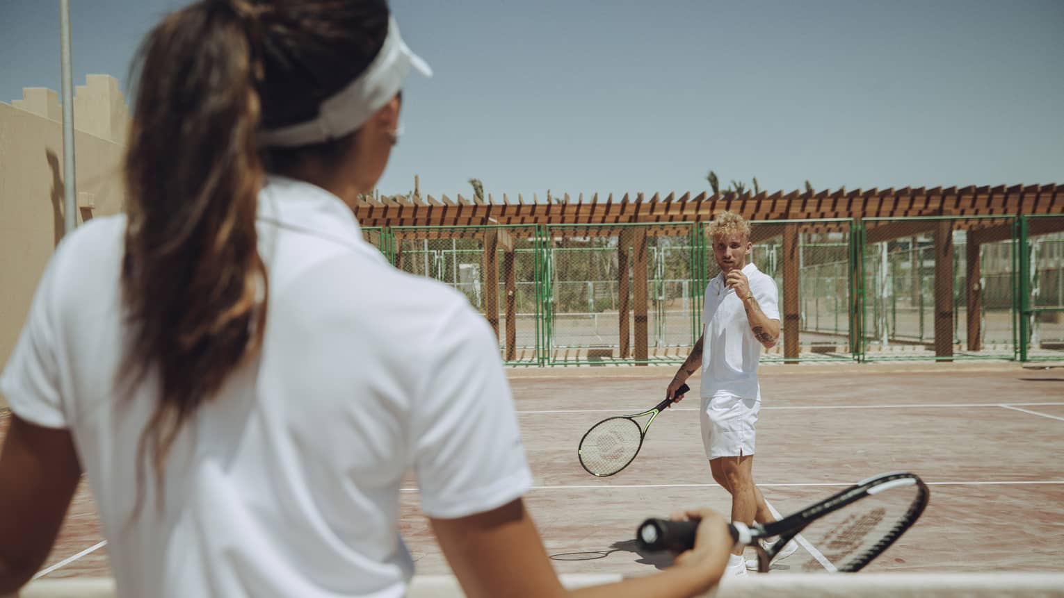 Two guests in tennis whites with rackets face each other on a court, one seen from behind as the other prepares to serve.