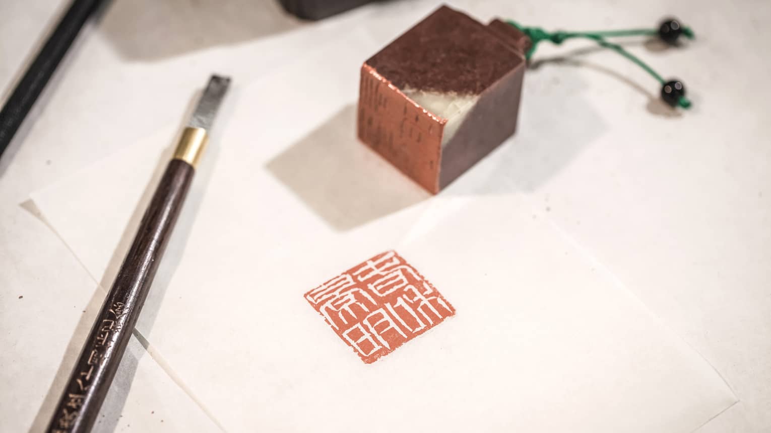 A small chisel and a little cube of dark red stone rest on a piece of paper which bears the red-ink stamp from a carved seal.