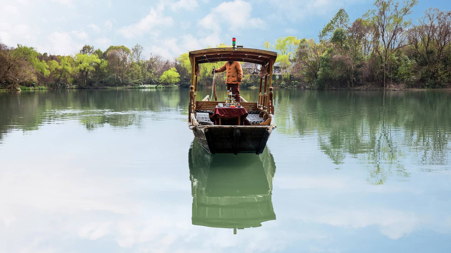 Reflected in the clear green water below, a forward-facing boat floats against a background of trees and blue sky.