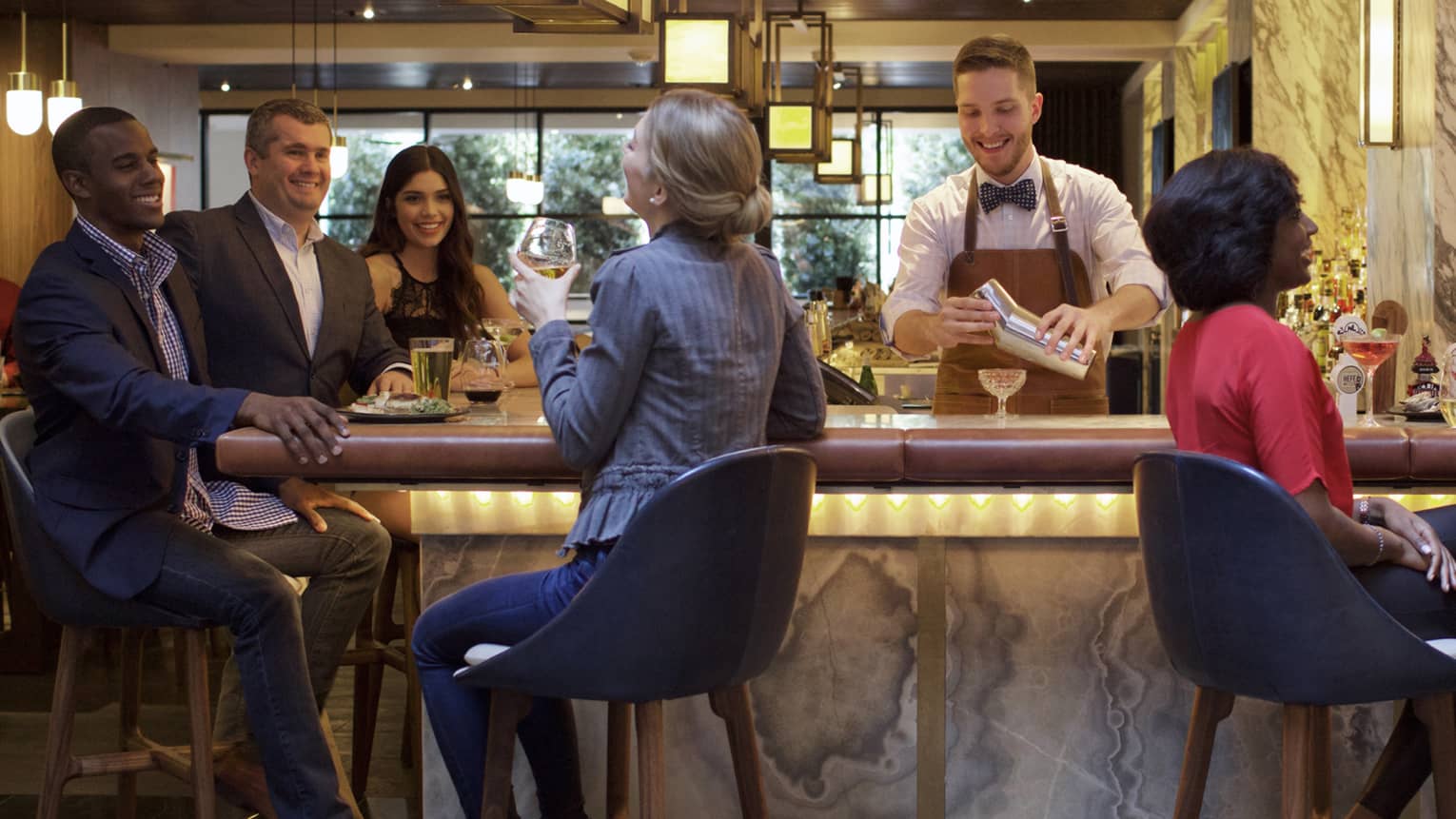 Two men and two women sitting on stools share a laugh at marble bar while smiling bartender pours cocktail from shaker 