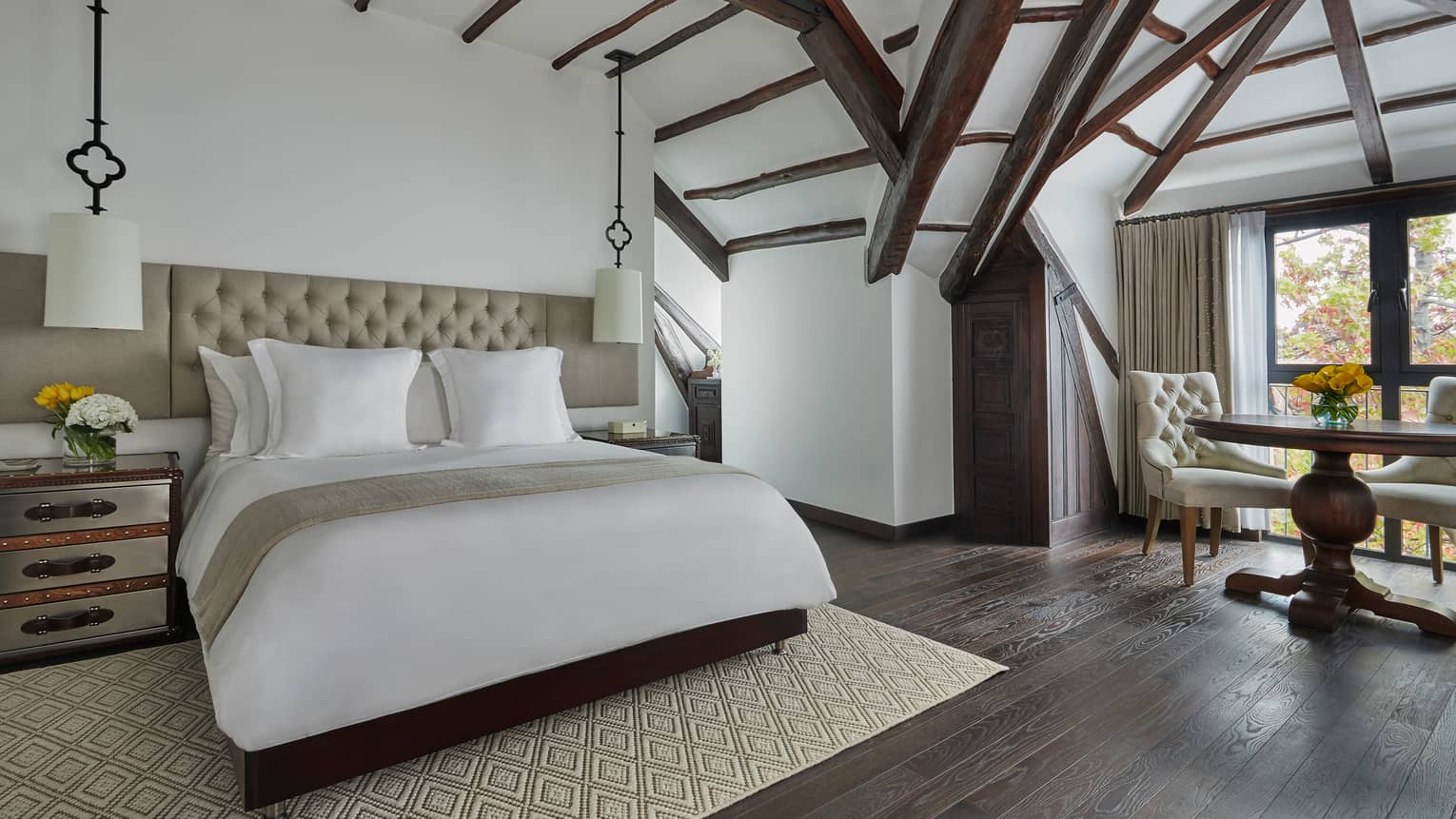 Beam ceiling in corner room over bright windows with table, chairs, bed with white linens, padded headboard, hanging lamps