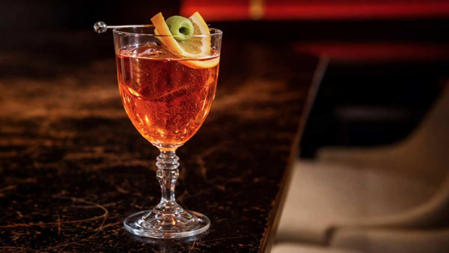 A clear, stemmed cocktail glass holds a clear reddish-orange cocktail with citrus slice and olive garnish