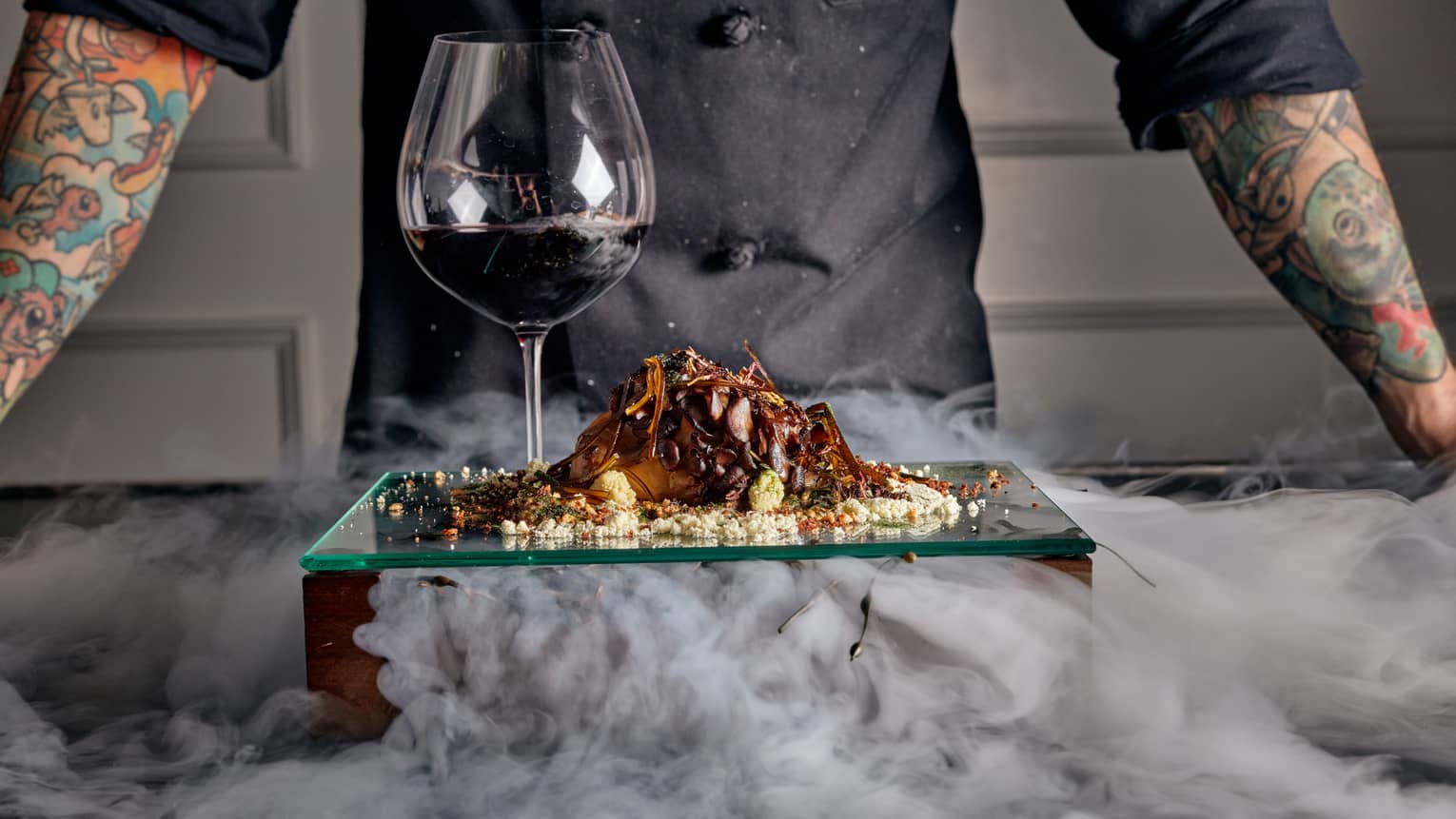 A chef standing behind a glass plate of food and a glass of red wine.