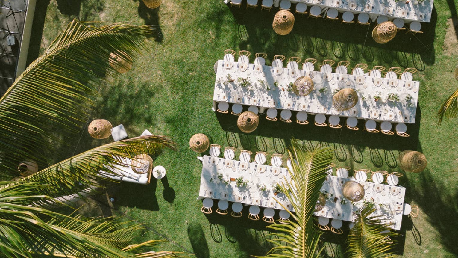 An aerial view of an outdoor event setup on a green lawn surrounded by palm trees. Setup includes long dining tables set for an event, chairs, lighting and other decor.