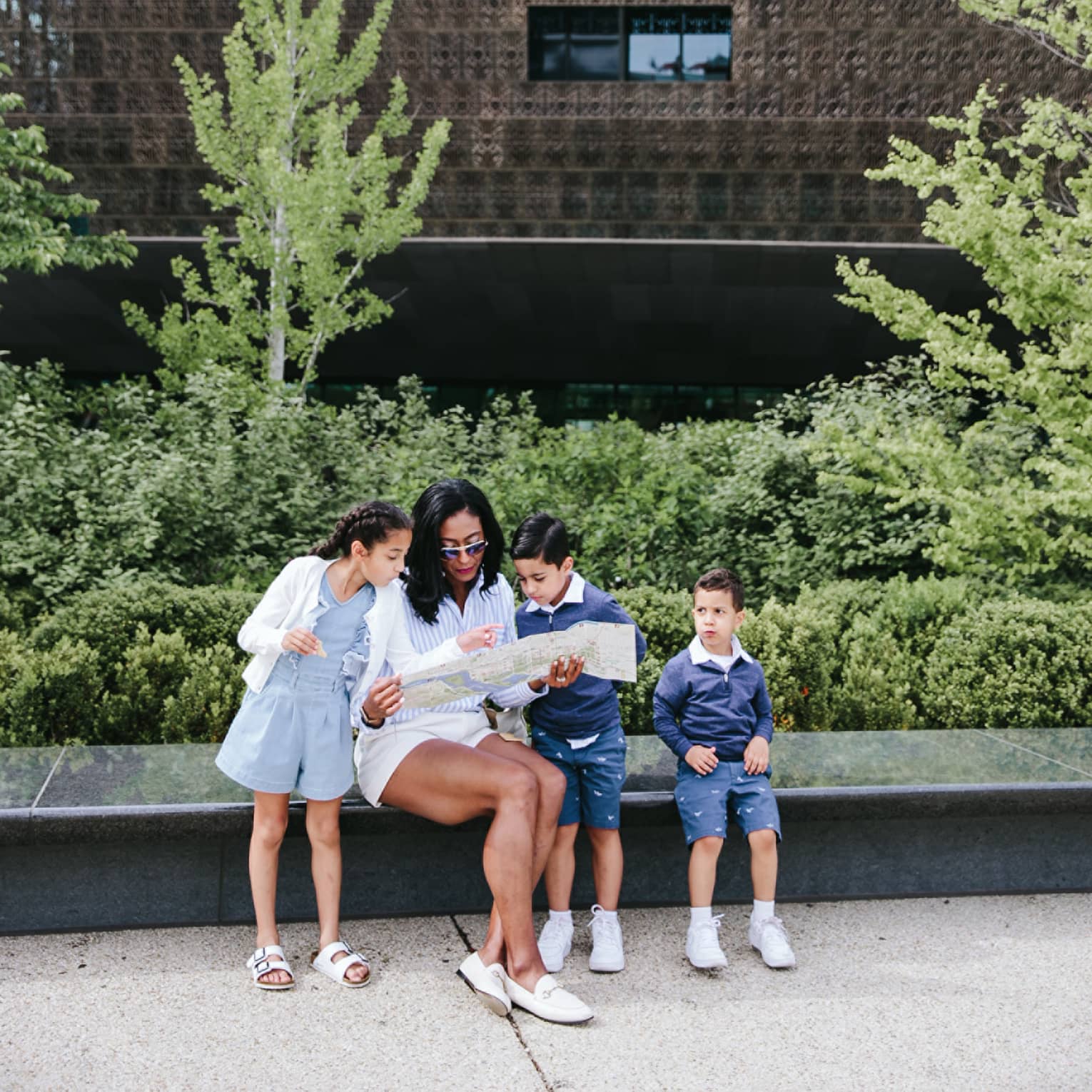 Woman and three young children sit on bench in city with landscaping behind them