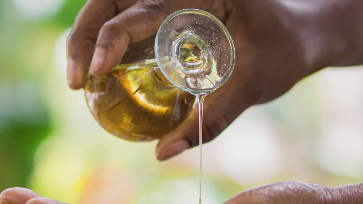 Golden oil flows from a small carafe held in one hand onto the other, a small wooden bowl underneath to catch the overflow.