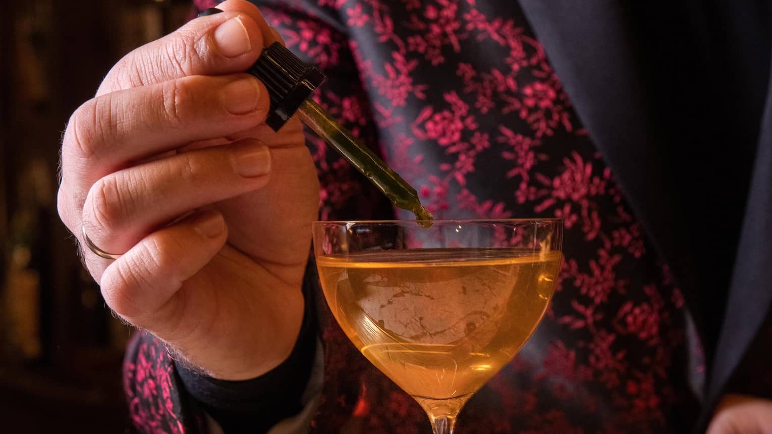 Bartender in red blazer uses dropper to add ingredient to bright-orange Basil cocktail in cordial glass
