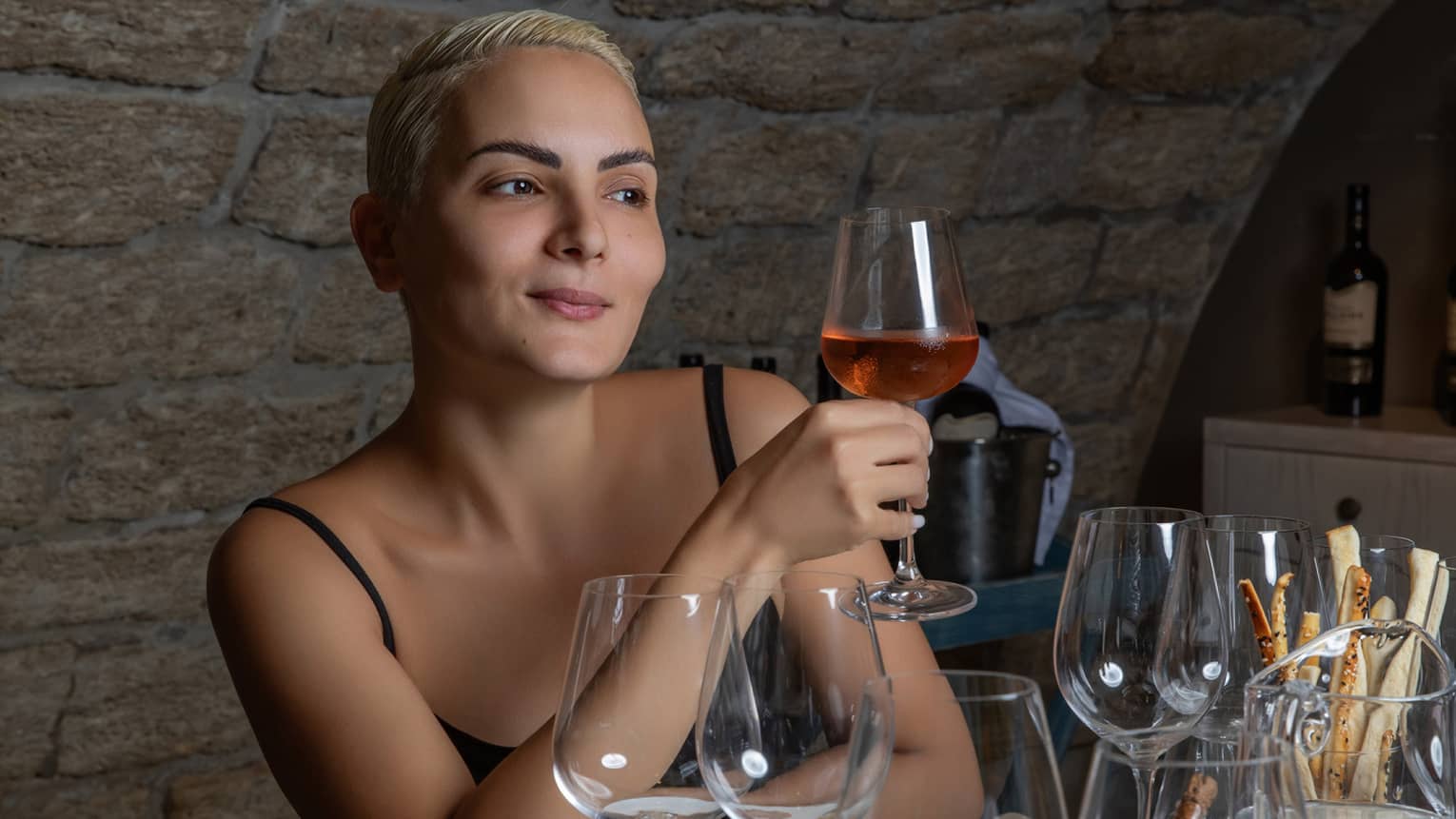 At a table set with glassware and hors-d’oeuvres, a wine enthusiast holds a glass containing a rose-coloured beverage.