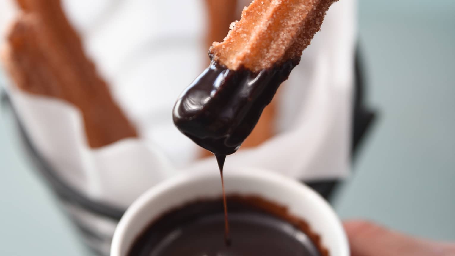 Hand holds small white bowl of melted chocolate, dipping churro into it