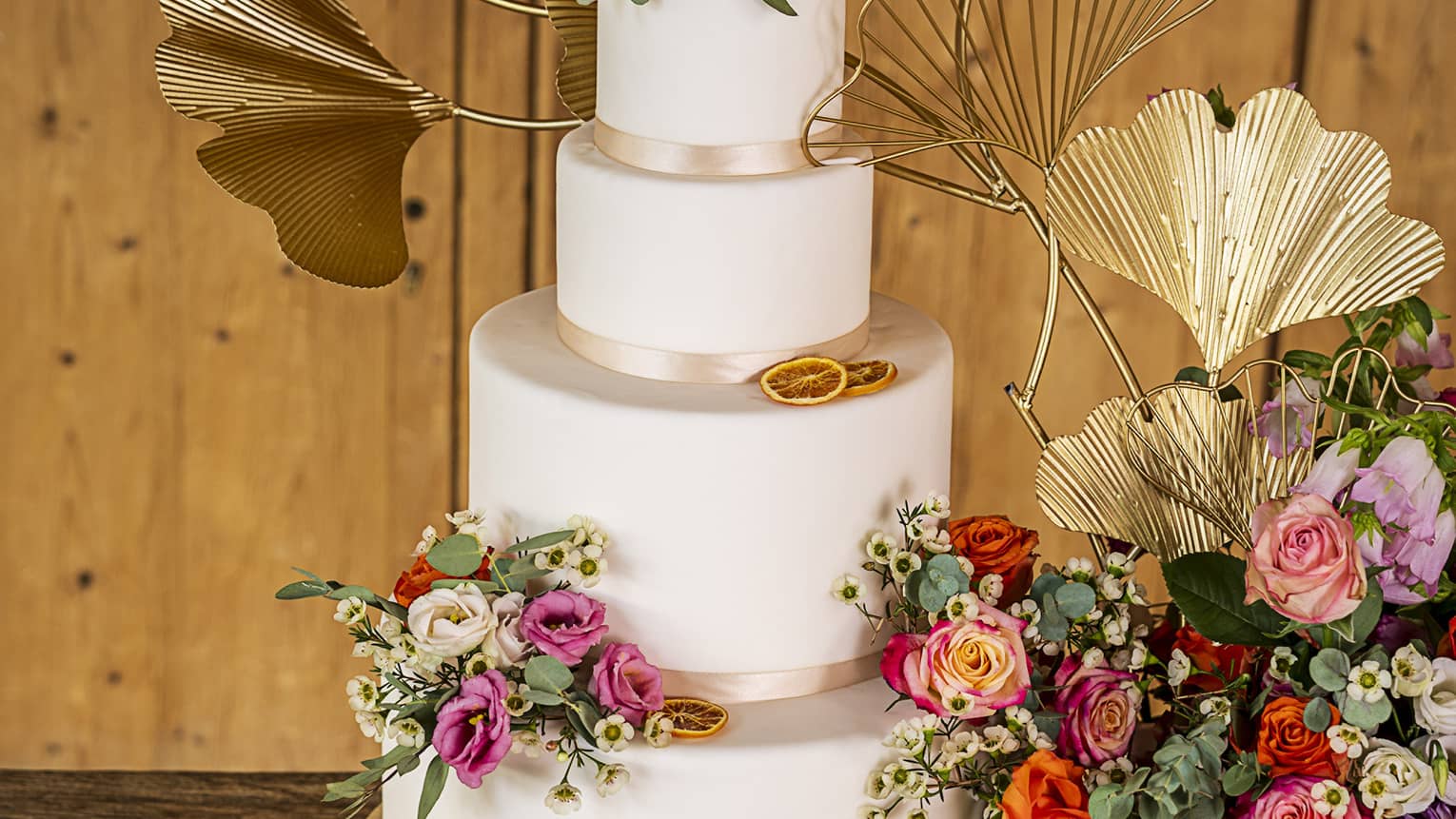 Three-tiered wedding cake with autumn-coloured flower and gold metal gingko accents beside large bouquet