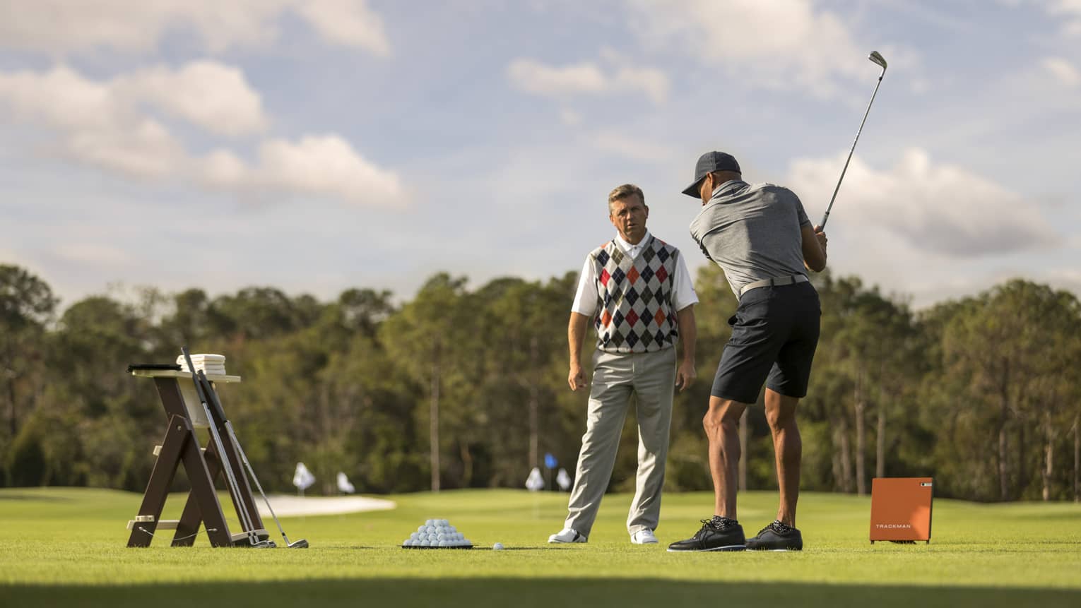 Against clear sky, one golfer looks on as another prepares to swing, a tray of balls and a stand of clubs nearby.