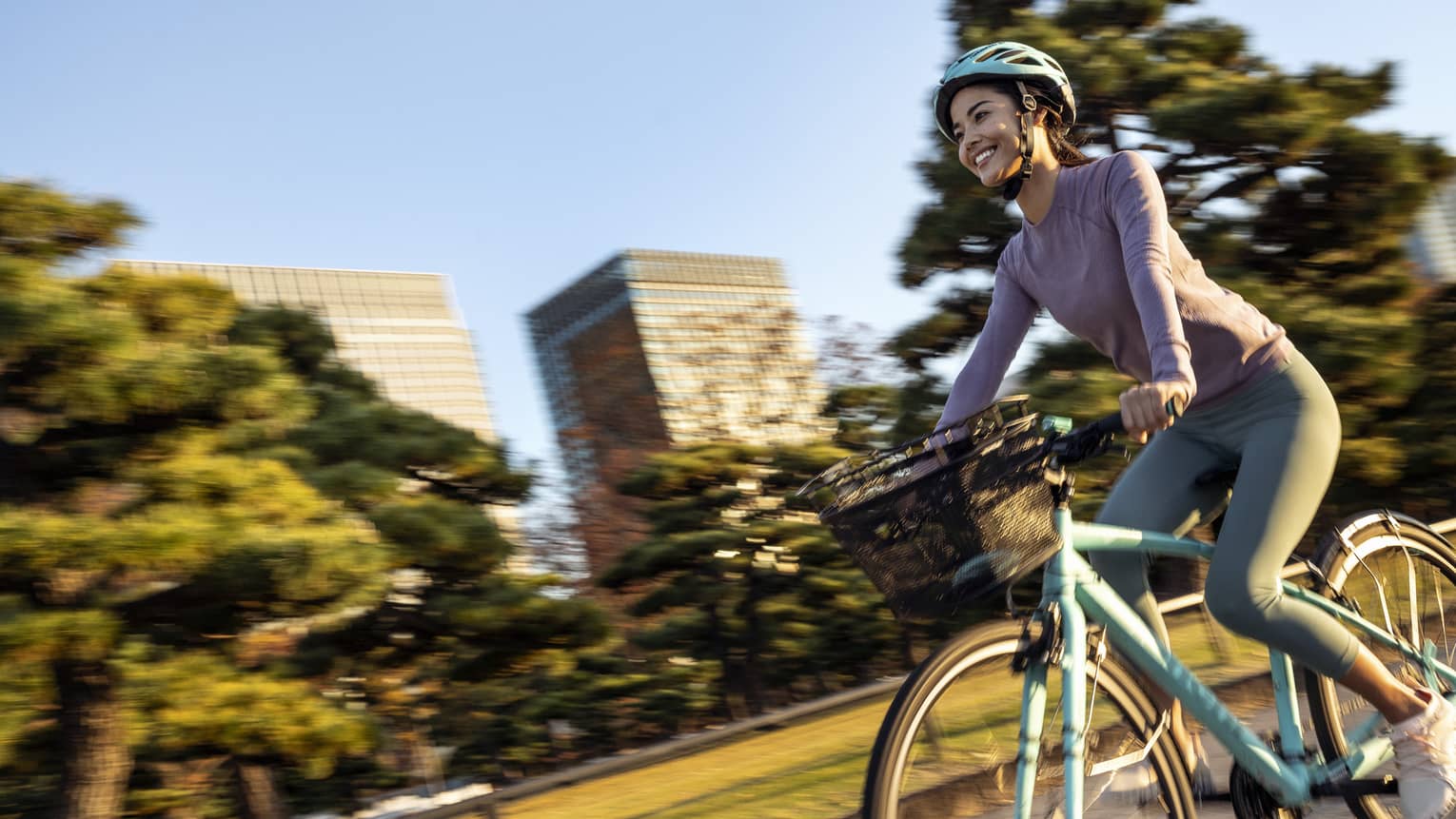 Smiling woman on teal cruiser bike with matching helmet rides by mature trees and skyscrapers of downtown Tokyo