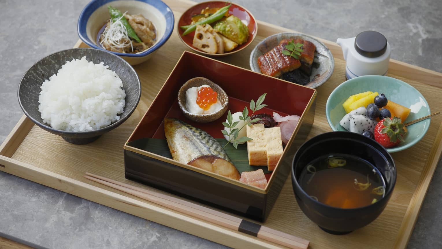 A close up of a Japanese breakfast on a natural wooden tray ? fish, chopsticks, rice, soup and fruit