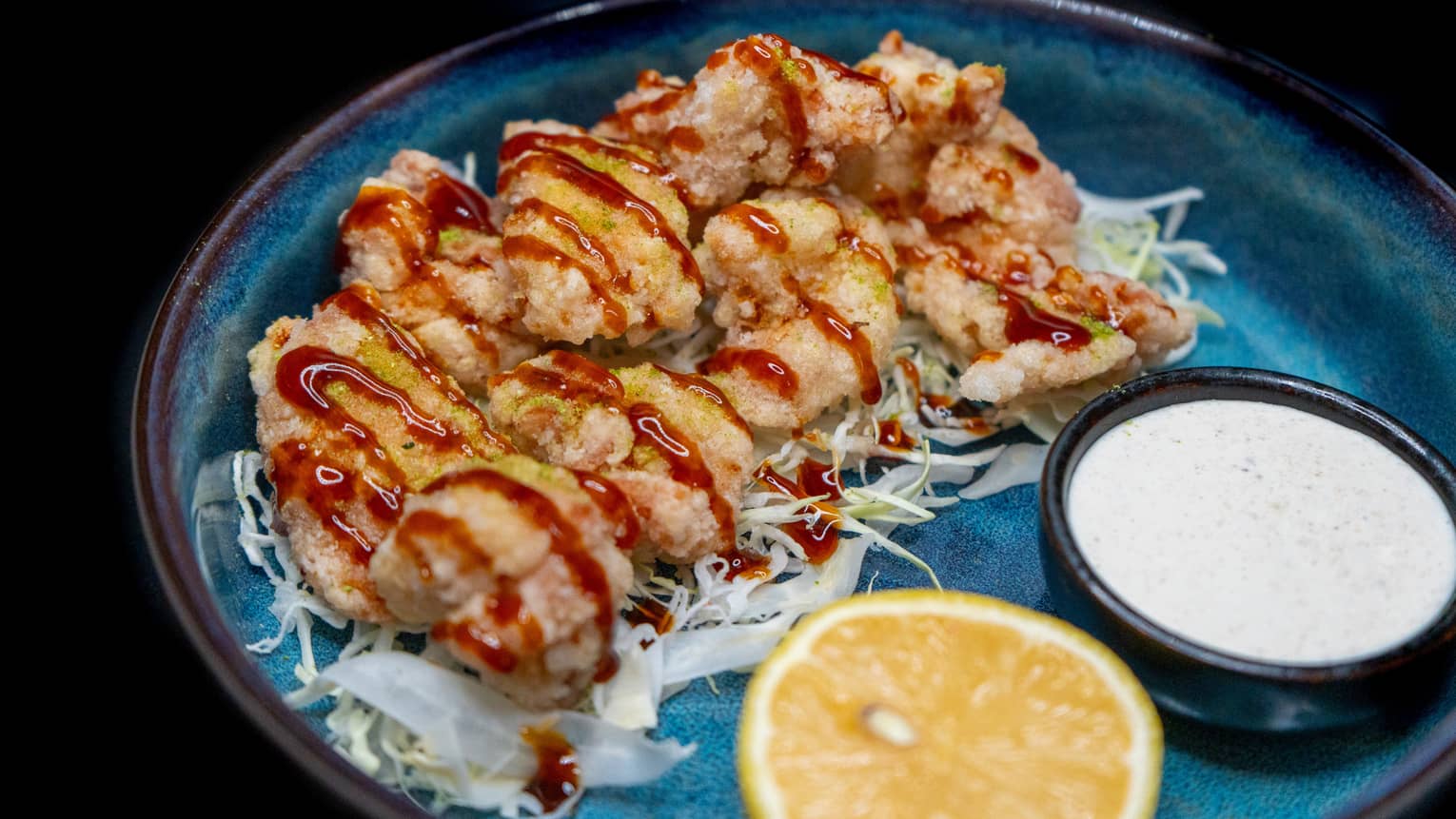 A blue plate holds light brown food items drizzled with a reddish sauce. A small dish of white sauce and a slice of citrus fruit garnish the plate.