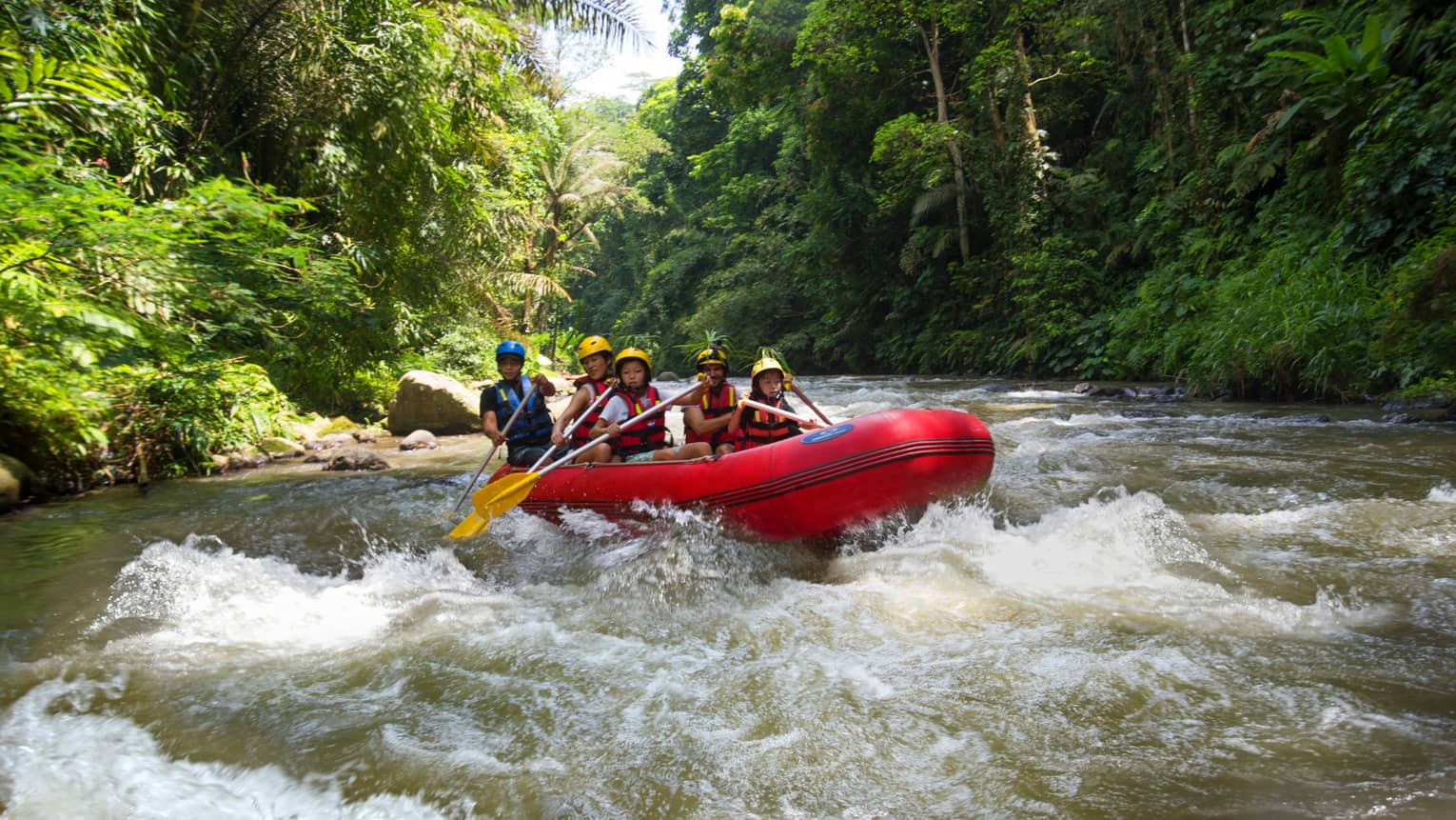 Family of four and guide hold paddles, steer red inflatable raft on rapids in river