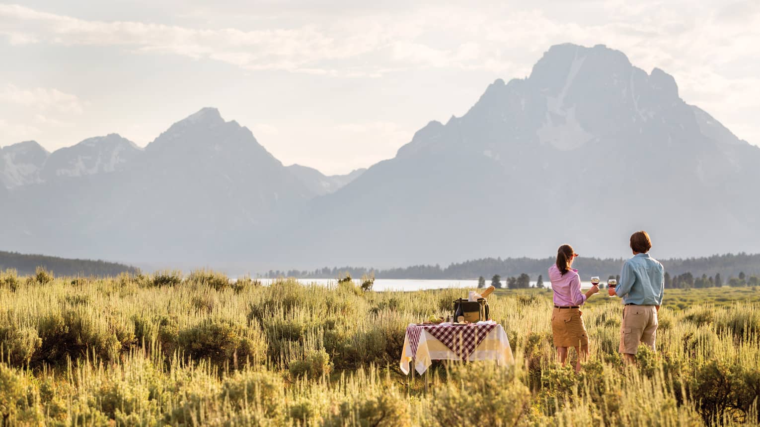 A man and woman sip wine in the grasslands while gazing out at the mountains at sunset