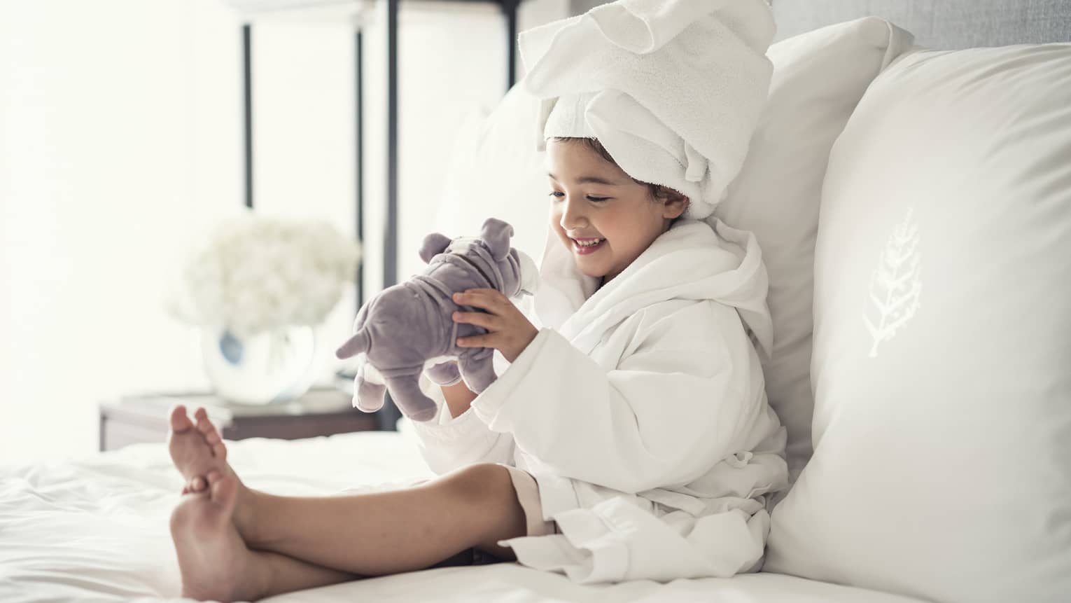 Young girl wearing a white bathrobe with towel wrap plays with plush dog toy on bed