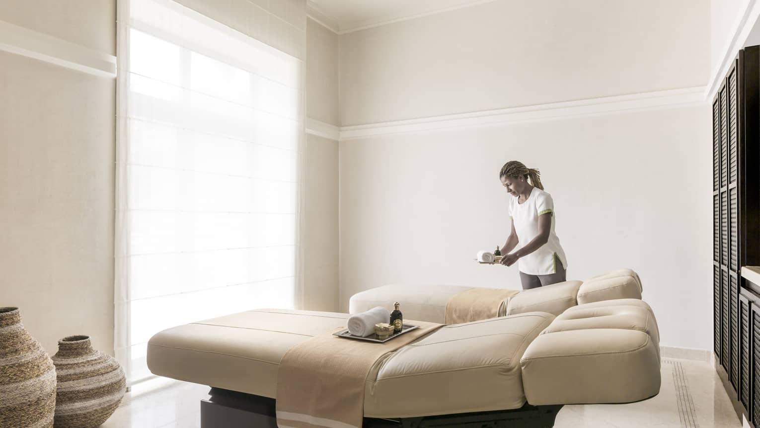 Hotel staff sets tray on Spa Couples Suite massage bed in bright treatment room