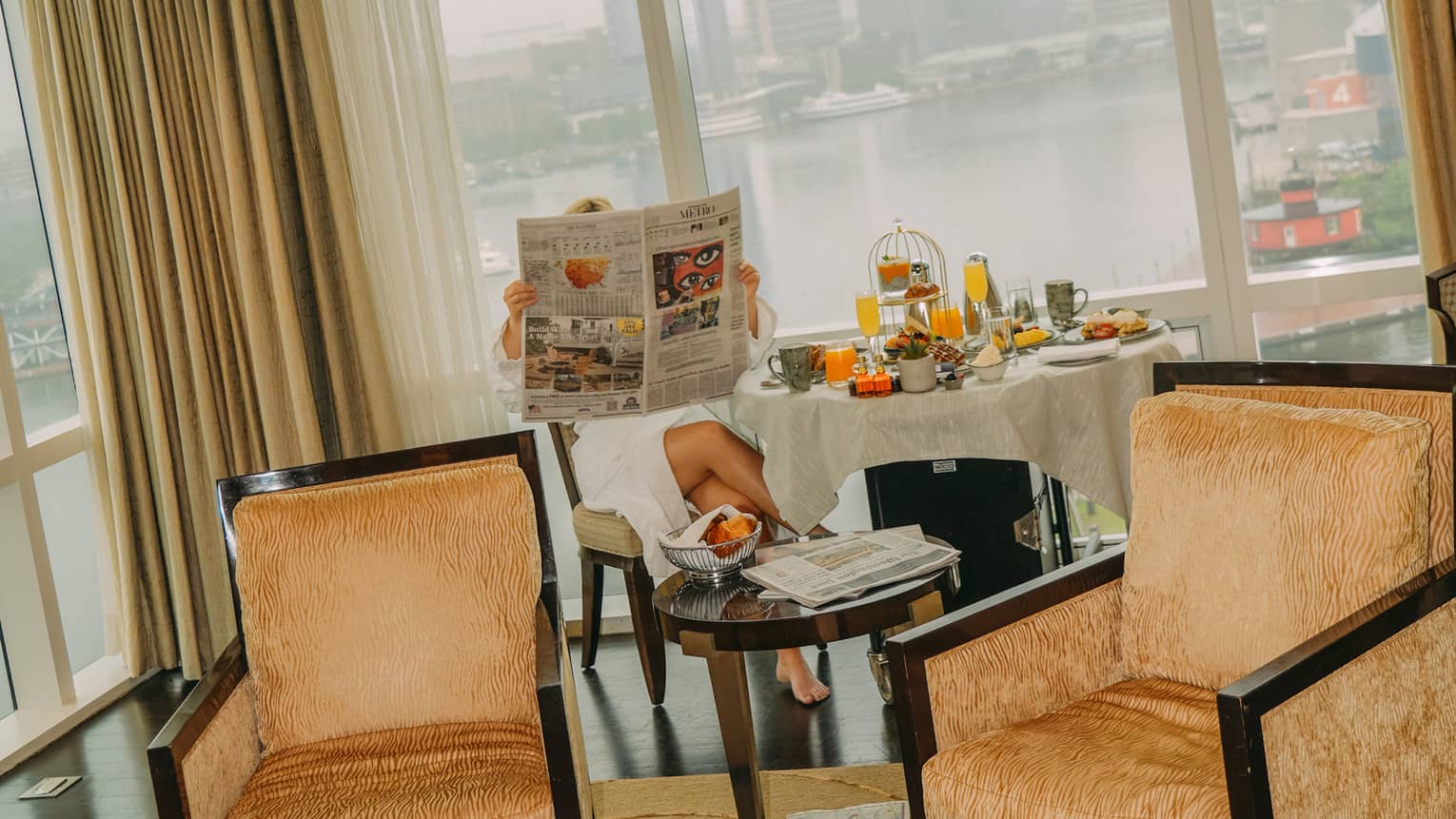 A woman dressed a white bathrobe reads a newspaper while seated at a dining table filled with breakfast food overlooking a harbor view