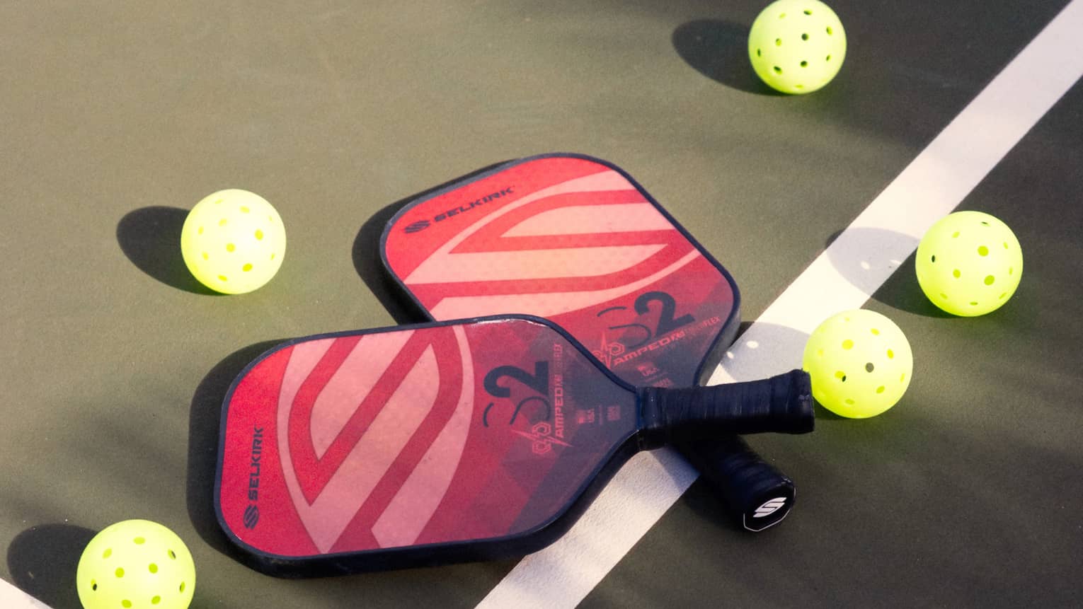 Close-up of two red and pink pickleball racquets, handles crisscrossed on a white court line, surrounded by five pickleballs.