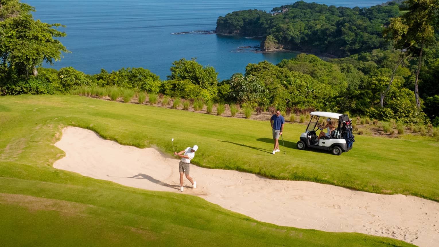 One golfer hits their ball out of a sand trap as another golfer stands by a golf cart looking on, with the ocean in the background
