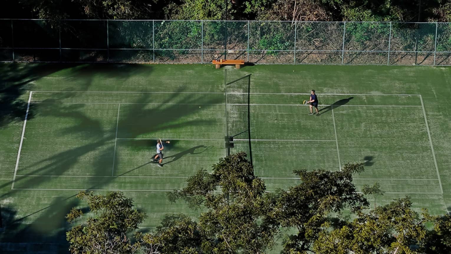 Two people play tennis on a green court surrounded by trees.