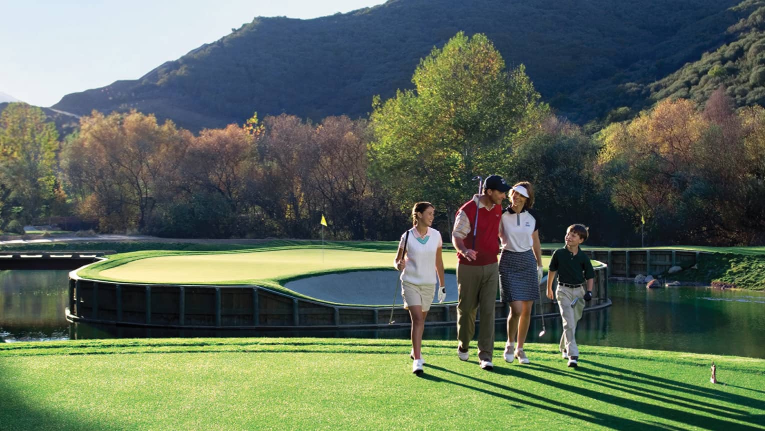 Family holds golf clubs, walks along sunny green on course near ponds, mountains