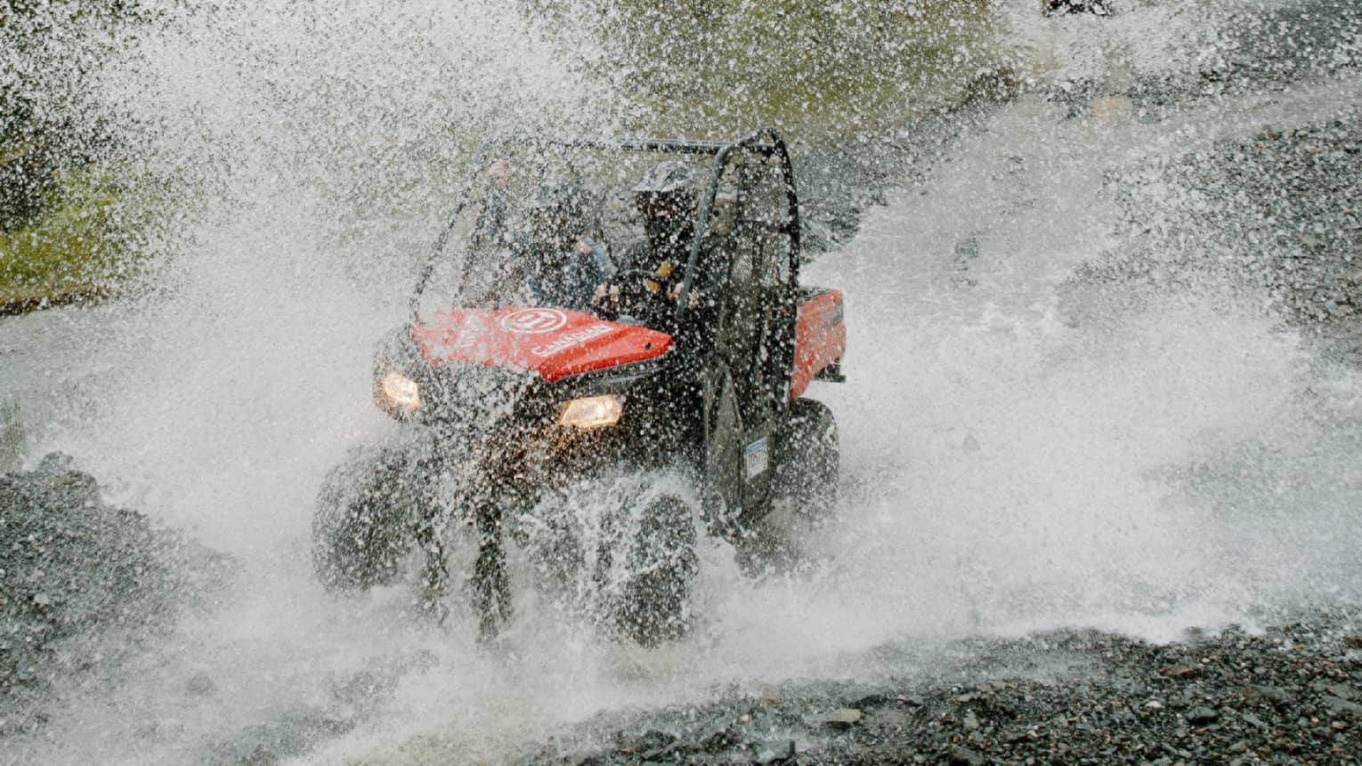 A red-and-black ATV driving through water, creating a thick veil of water spray against a background of pine forest.