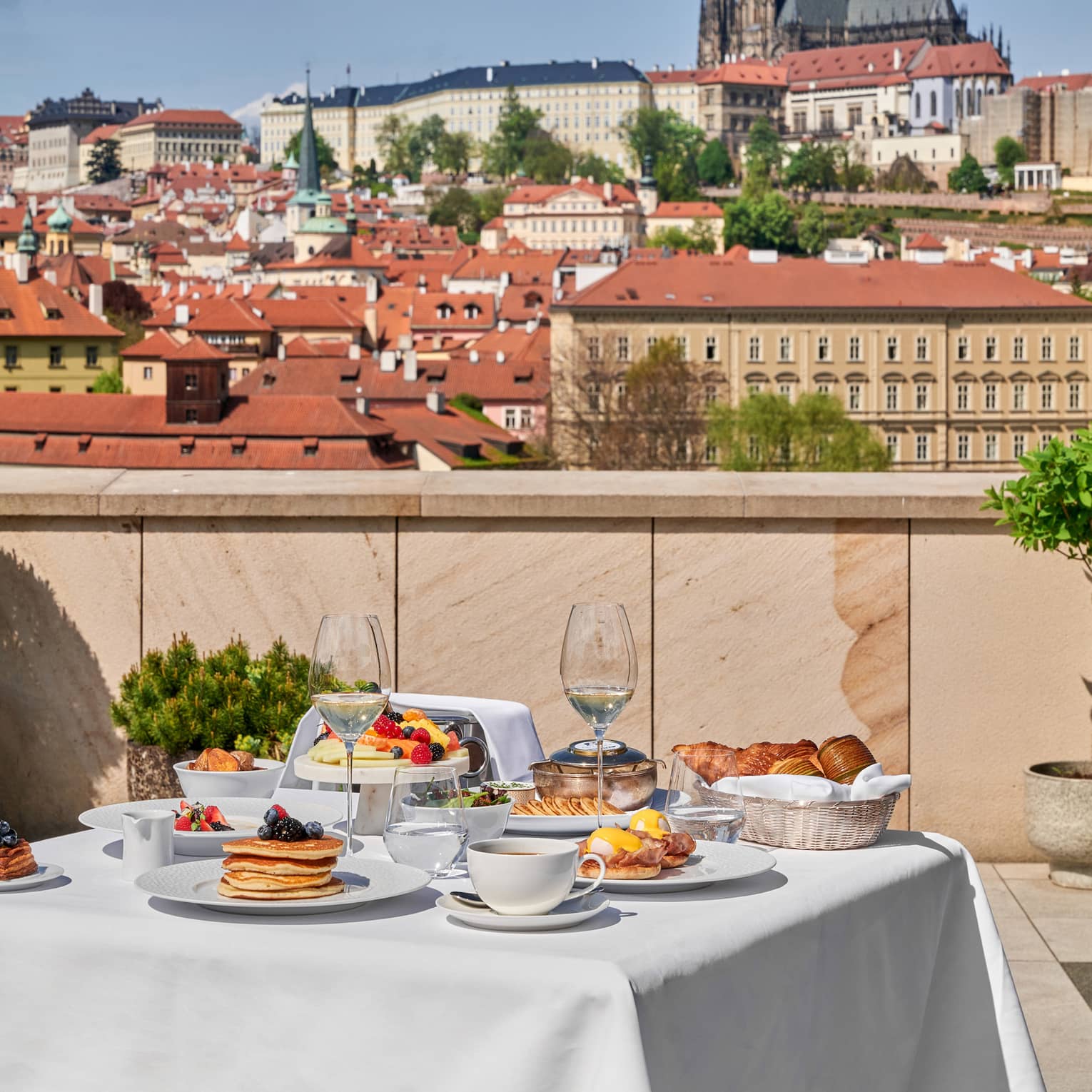 A white cloth?covered dining table on a terrace, set for a meal, with city views in the background