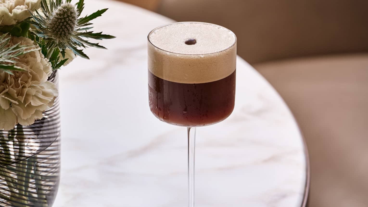 An espresso cocktail sits atop a white marble tabletop next to a floral arrangement