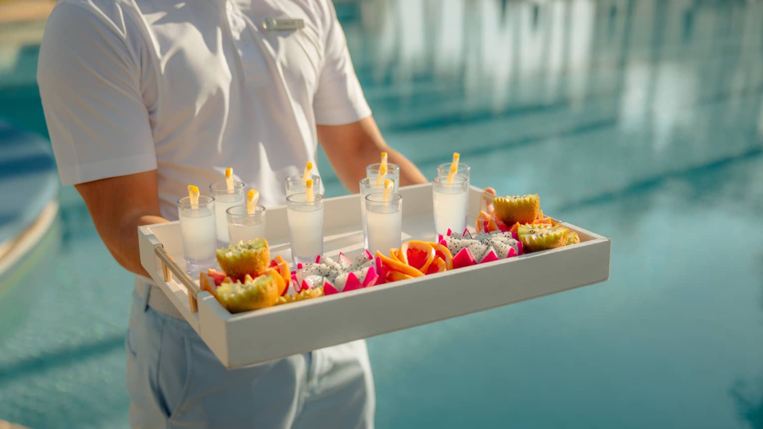 Pool attendant wearing a white short-sleeve polo shirt and light-blue shorts carries a large tray filled with beverages and small plates of colourful fruit and snacks