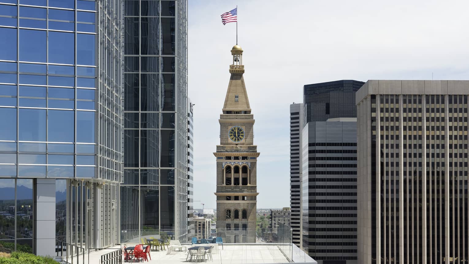 Denver's distinctive, historic clock tower, American flag on top, rises above a top floor patio amid modern skyscrapers.