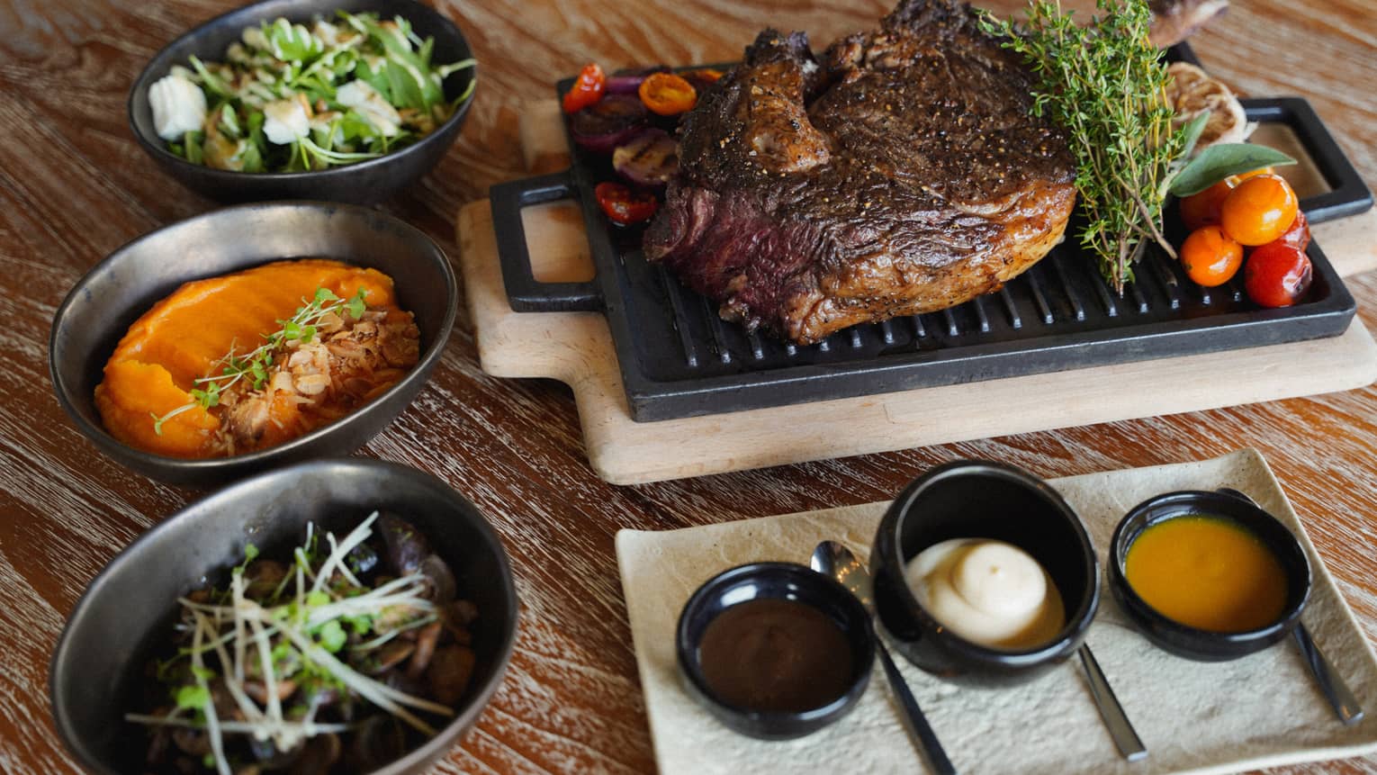 A serving platter on a wood table holds a piece of cooked meat surrounded by garnishes. Three small oval dishes next to the platter hold colorful sides, and a smaller platter holds three small bowls containing condiments.