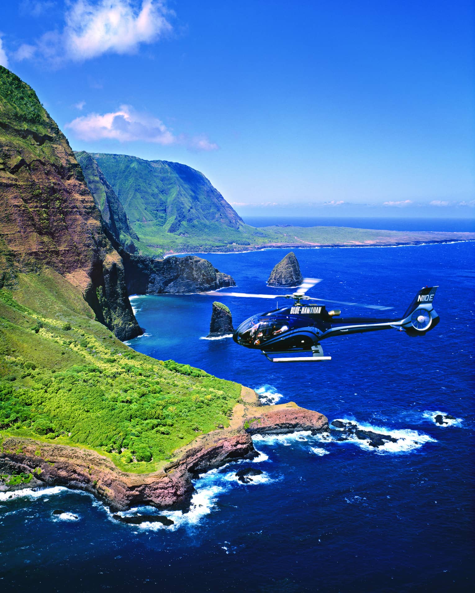 Helicopter flies over ocean and sweeping green cliffs 