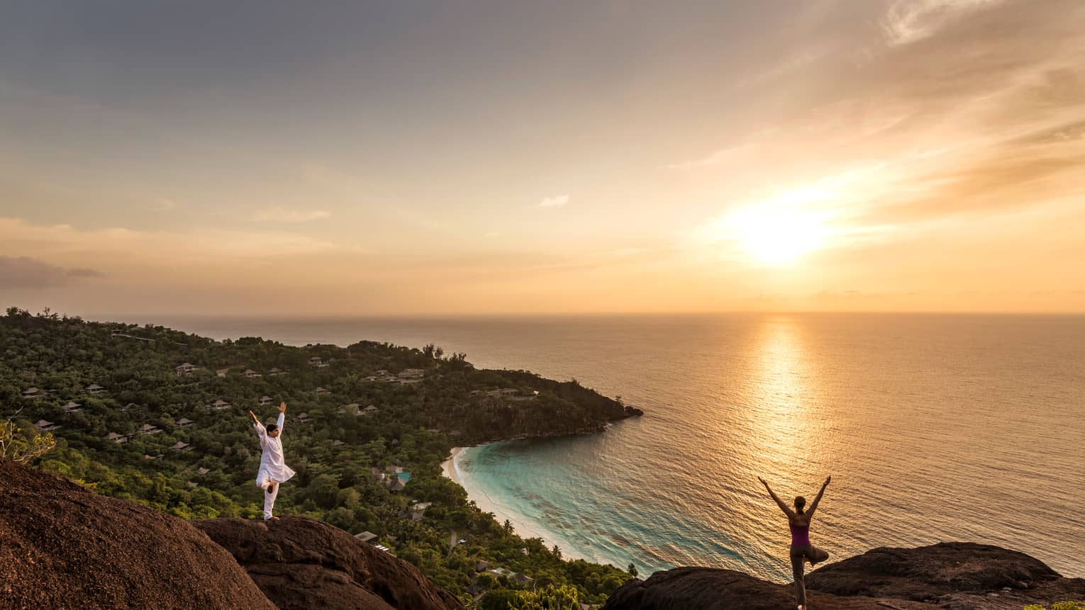 Woman and man stand apart on large boulders overlooking canopy of trees, ocean at sunrise 
